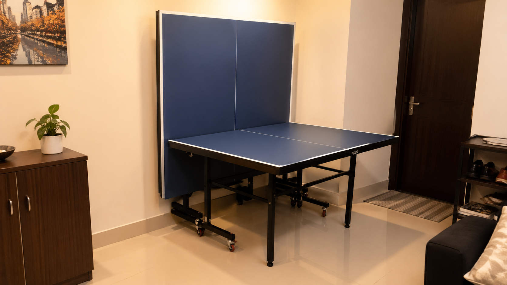 Foldable table tennis table in playback mode inside a compact Indian apartment, with one half folded upright for solo practice and caster wheels visible on the sturdy black frame